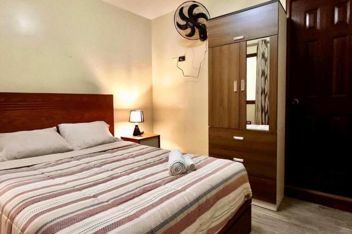 Quarto de hotel com cama, mesa de cabeceira, luminária, espelho e guarda-roupa.
