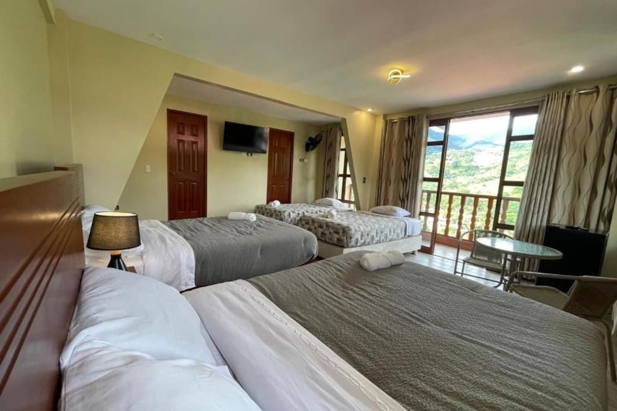 Quarto de hotel com duas camas, uma mesinha, televisão e varanda com vista.