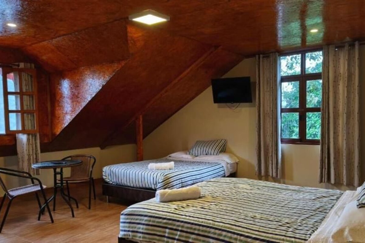 Quarto com teto inclinado, cama com roupa de cama listrada, uma pequena mesa com cadeiras e televisão fixada na parede.
