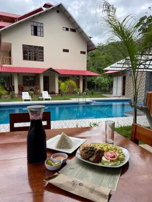 Uma mesa de jantar com uma refeição, uma piscina e uma casa ao fundo.