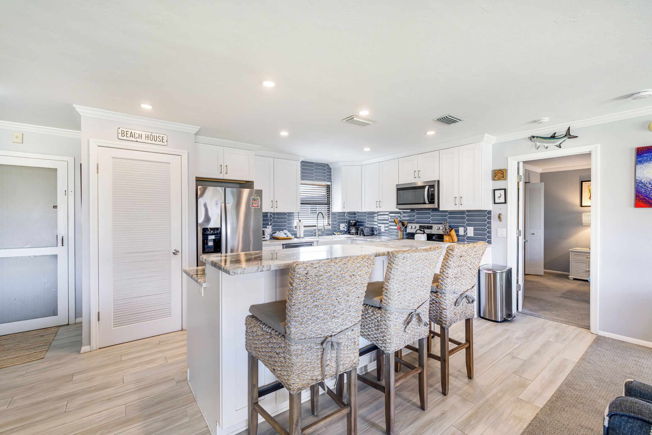 A modern kitchen with white cabinets, a central island with bar stools, and a sign reading 'BEACH HOUSE'.