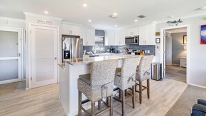 Una cocina moderna con armarios blancos, una isla central con taburetes y un letrero que dice 'BEACH HOUSE'.