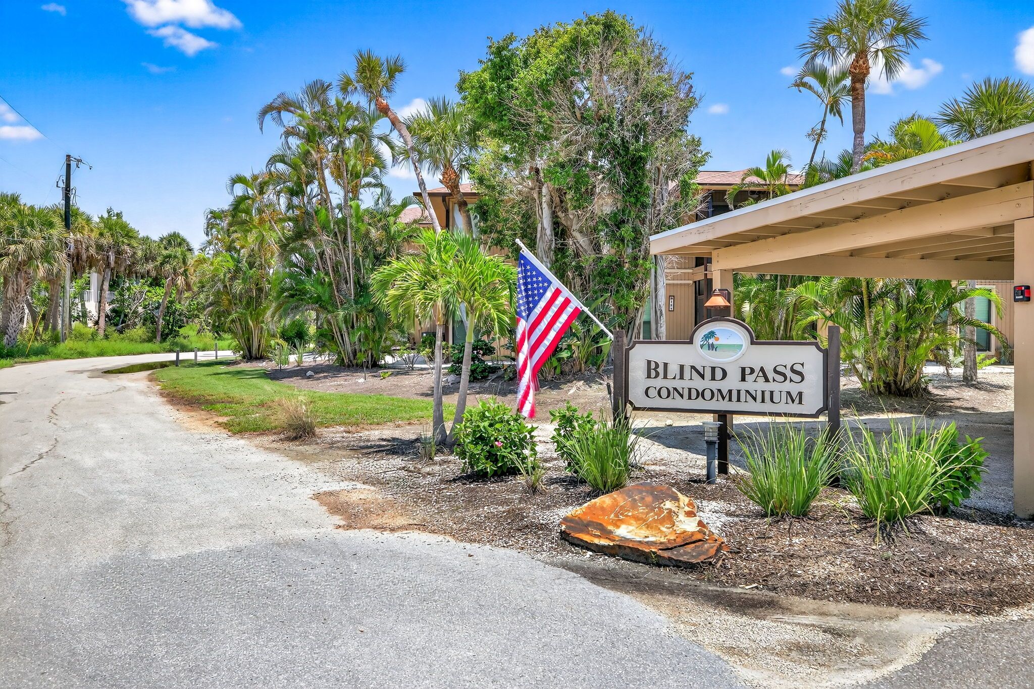A sign for a condominium with an American flag, surrounded by palm trees and a building with a covered entrance.