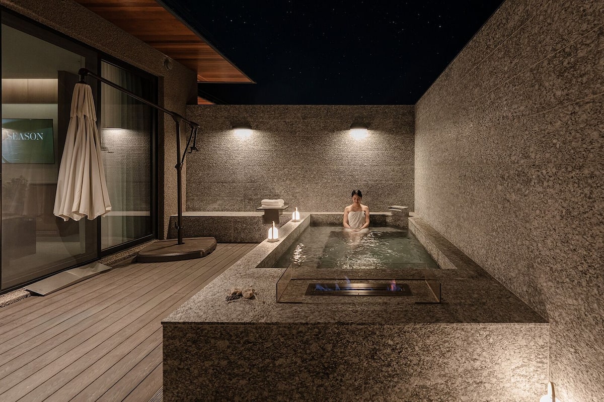 Un bain à remous extérieur, entouré de murs en pierre, éclairé la nuit, avec une personne assise à l’intérieur, un banc et un porte-serviettes.
