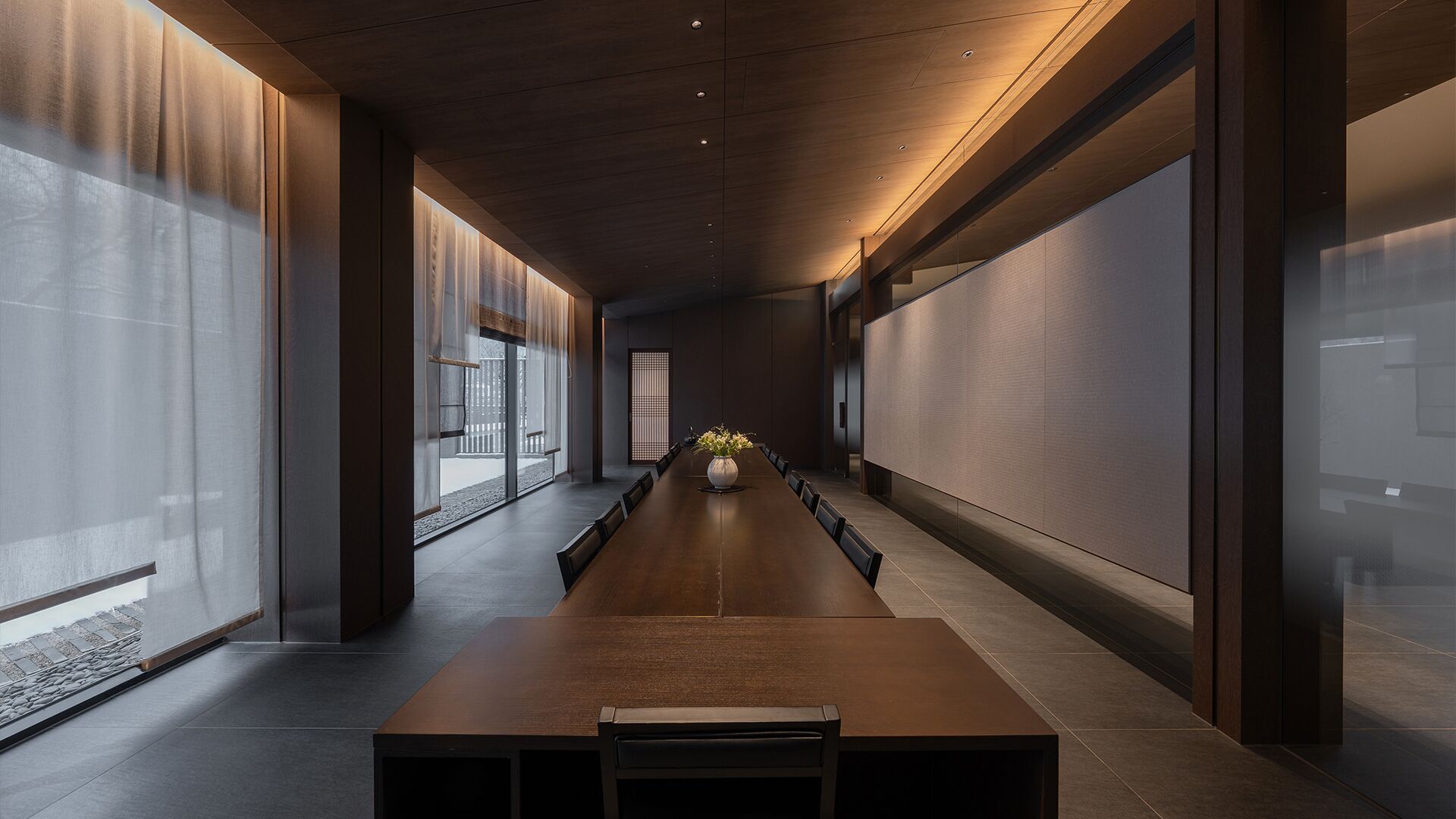 A long conference room with a wooden table and chairs, large windows, and a decorative vase.