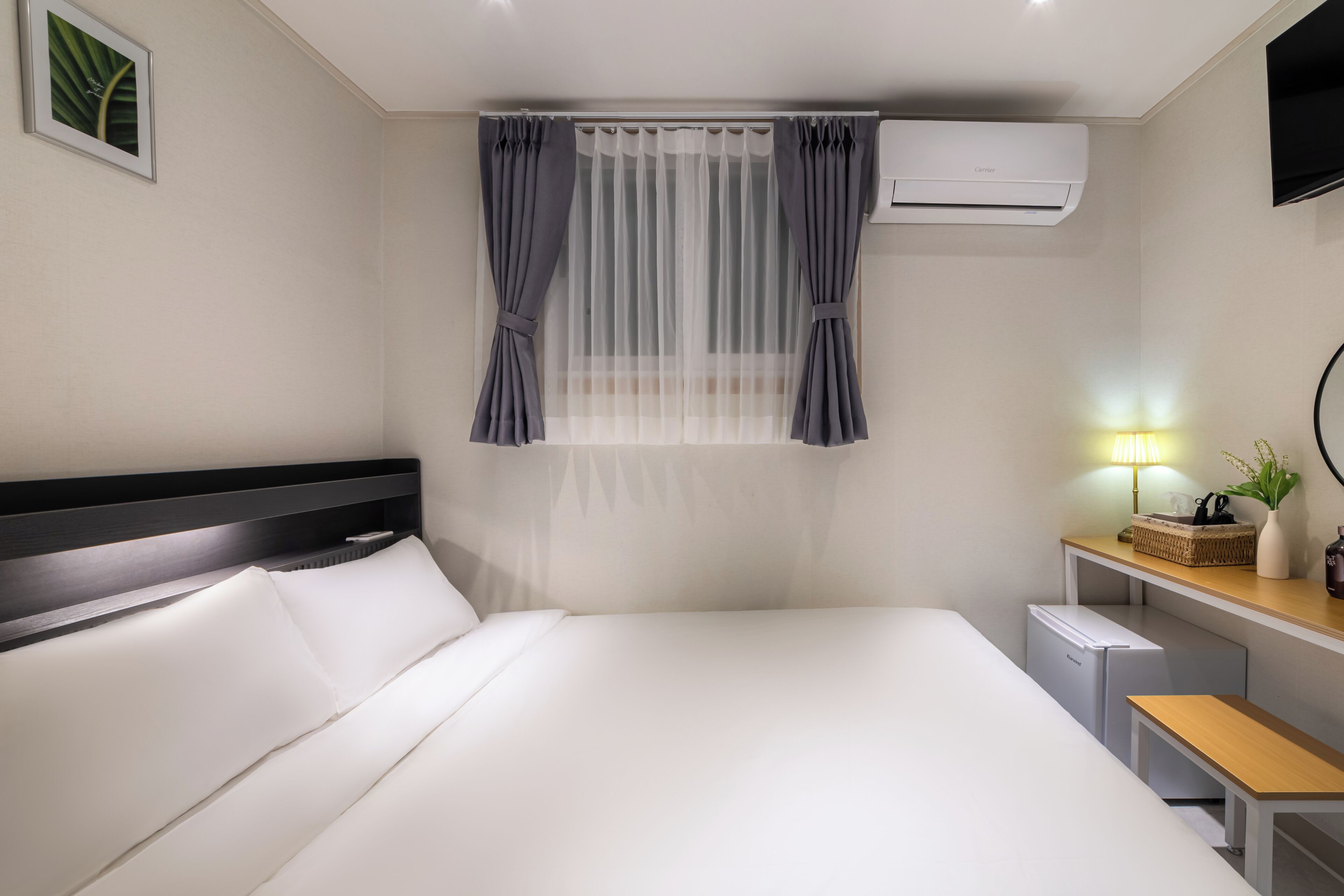 Une chambre d’hôtel équipée d’un lit, d’un bureau, d’un petit réfrigérateur, d’un climatiseur et d’une fenêtre avec des rideaux.