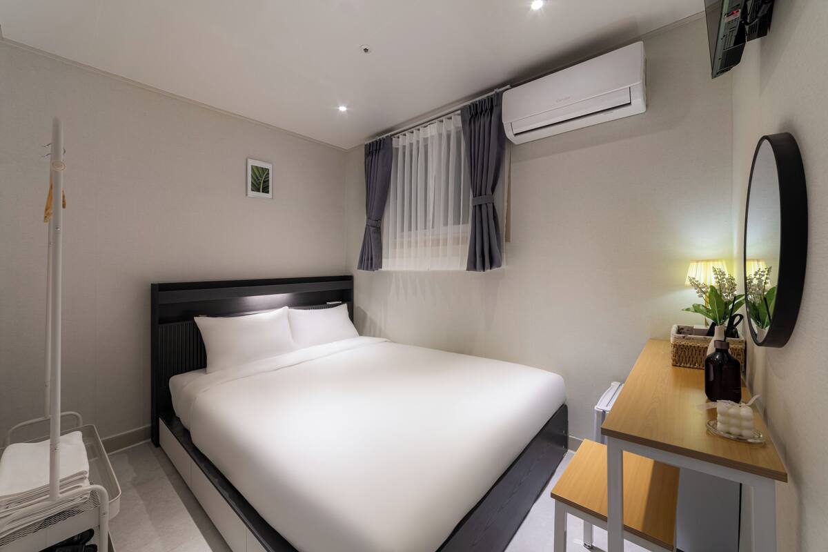 A hotel room with a bed, bedside table, mirror, air conditioning unit, and a small window with curtains.