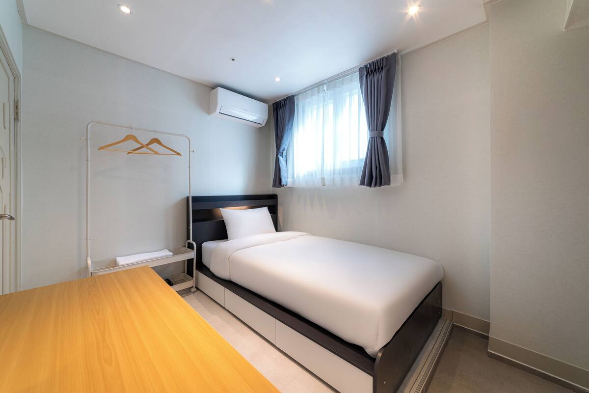 A single bed with a white mattress and headboard, a wooden table, a wall-mounted air conditioner, and a window with curtains.
