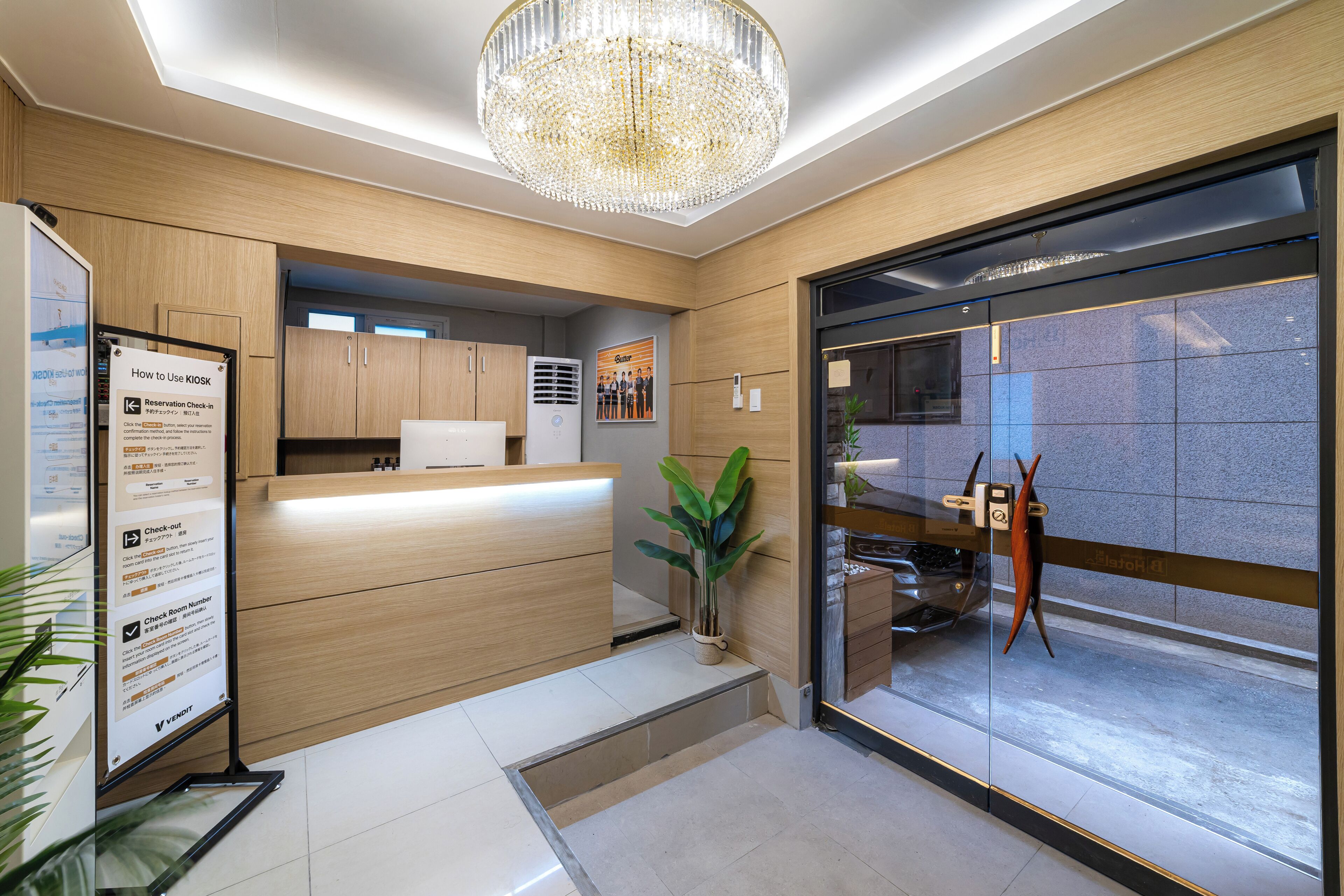 A reception area with a wooden reception desk, a large chandelier, a potted plant, and a glass door leading to a parking area.