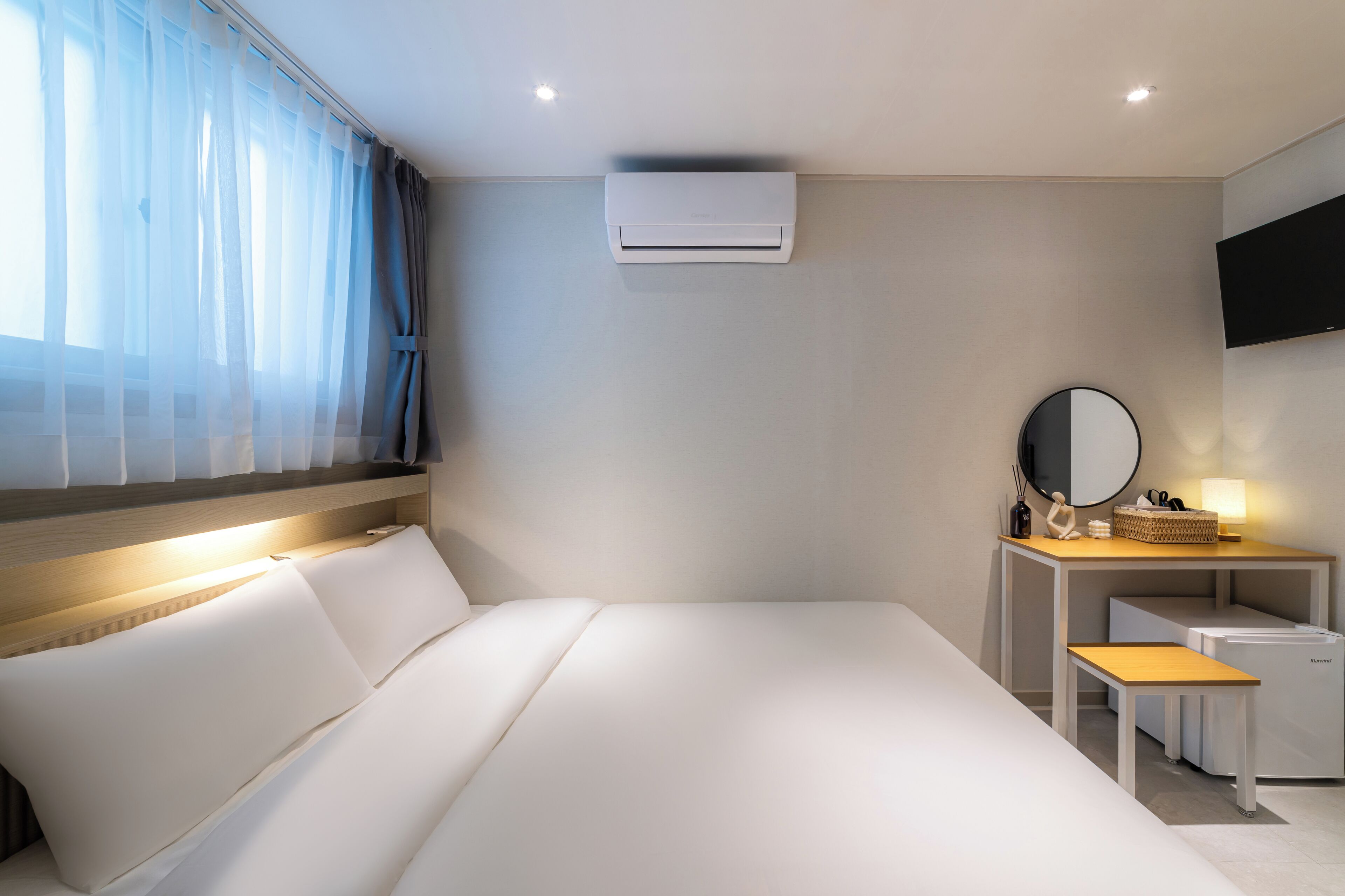 Hotellihuone, jossa on sänky, peilillä varustettu työpöytä, pieni pöytä, ilmastointilaite ja seinälle asennettu televisio.