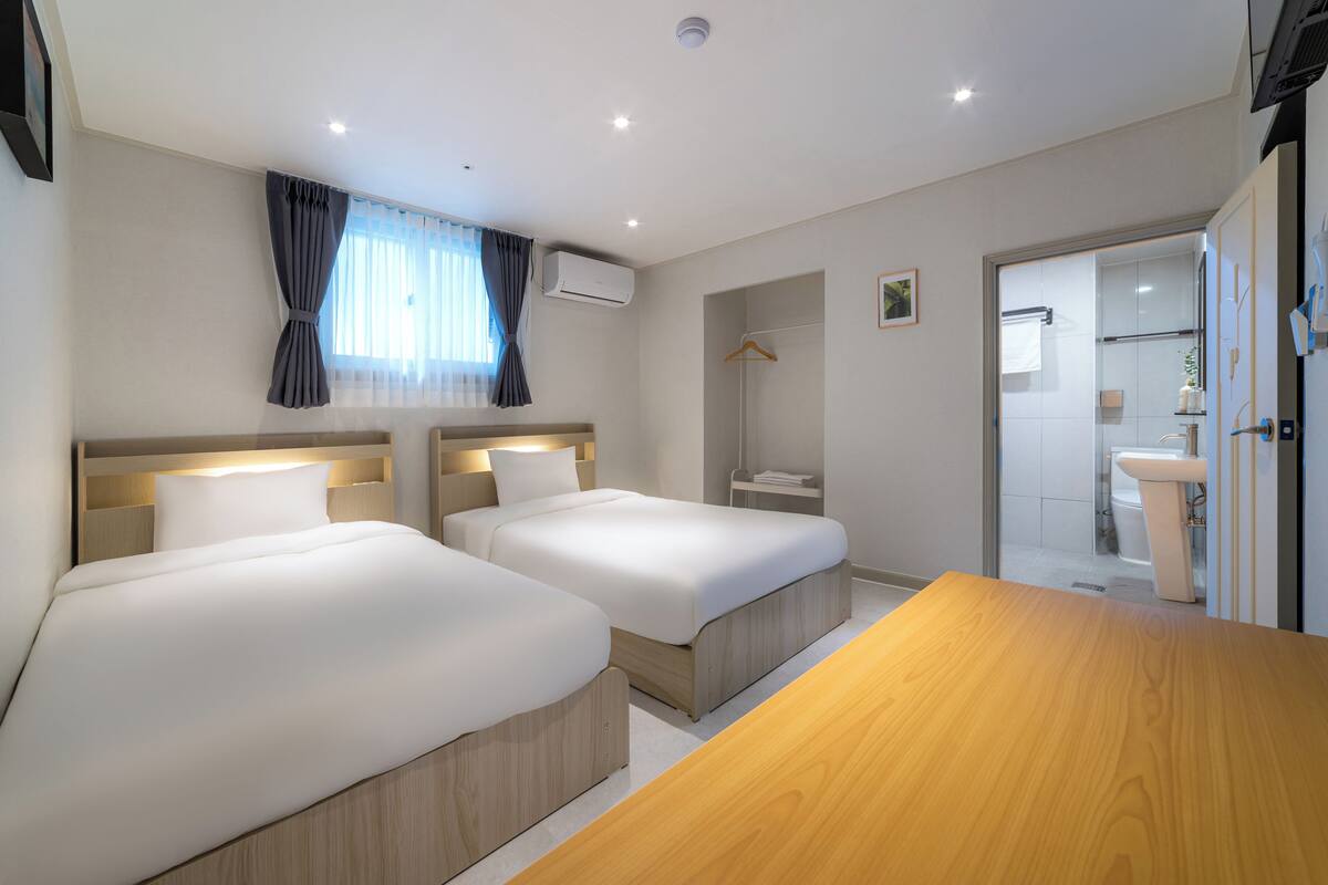 A hotel room with two beds, a wooden table, a bathroom with a sink and toilet, and a window with curtains.