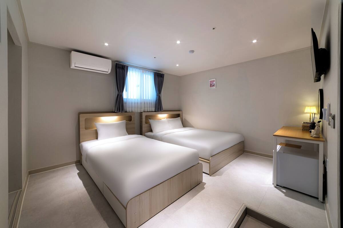 A hotel room with two beds, a desk, a TV, and an air conditioning unit.