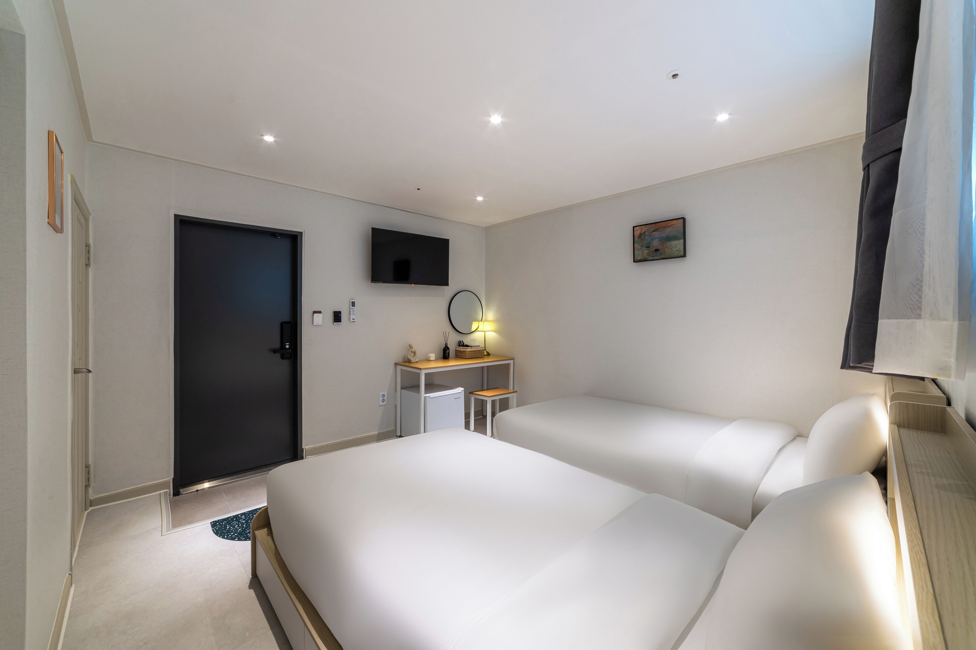 Moderni hotellihuone, jossa on kaksi sänkyä, taulutelevisio, työpöytä lampun kanssa ja pieni jääkaappi.