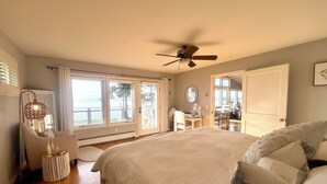 A bedroom with a bed, a ceiling fan, a desk with a chair, and a view of the outdoors through large windows. - Private Beach Front Executive Home (Bellingham)