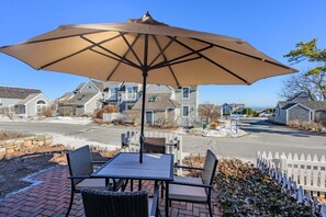 A patio with a table and chairs, an umbrella, and a view of houses in the background. - Blue Tide Retreat Steps from Private Beach (Mashpee)