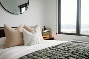 Een bed met een grijze deken, een ronde spiegel en een nachtkastje met een lamp en fotolijst.