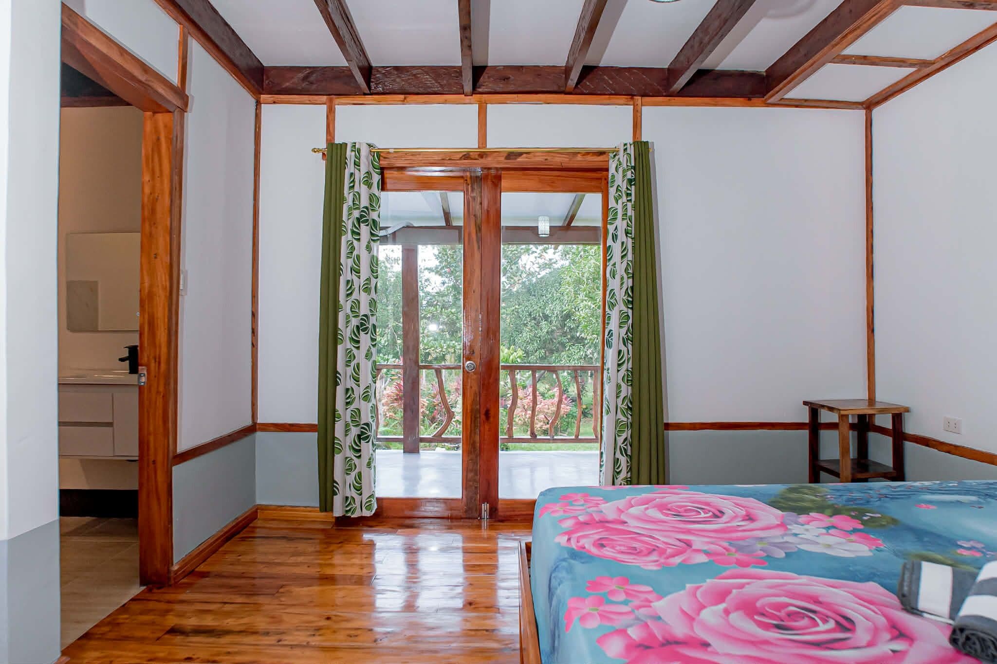 A room with a wooden floor, a bed with a floral bedspread, a wooden door with green curtains, and a view of greenery outside.