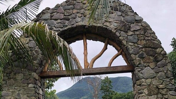 Ein steinernes Torbogen führt zu einem Gebäude mit weißem Dach, umgeben von Grünflächen und einer Berglandschaft im Hintergrund.