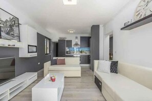 Ein modernes Wohnzimmer mit Sofa, Couchtisch und Fernseher. Im Hintergrund ist eine Küche zu sehen.