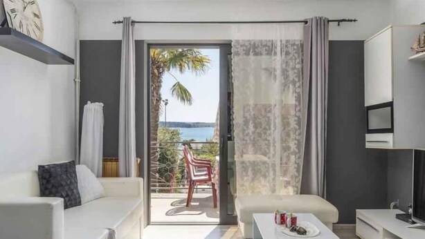 A modern living room with a sofa, coffee table, and TV stand. There is a sliding glass door leading to a balcony with a view of the sea. - Holiday apartment with air conditioning (Novigrad)