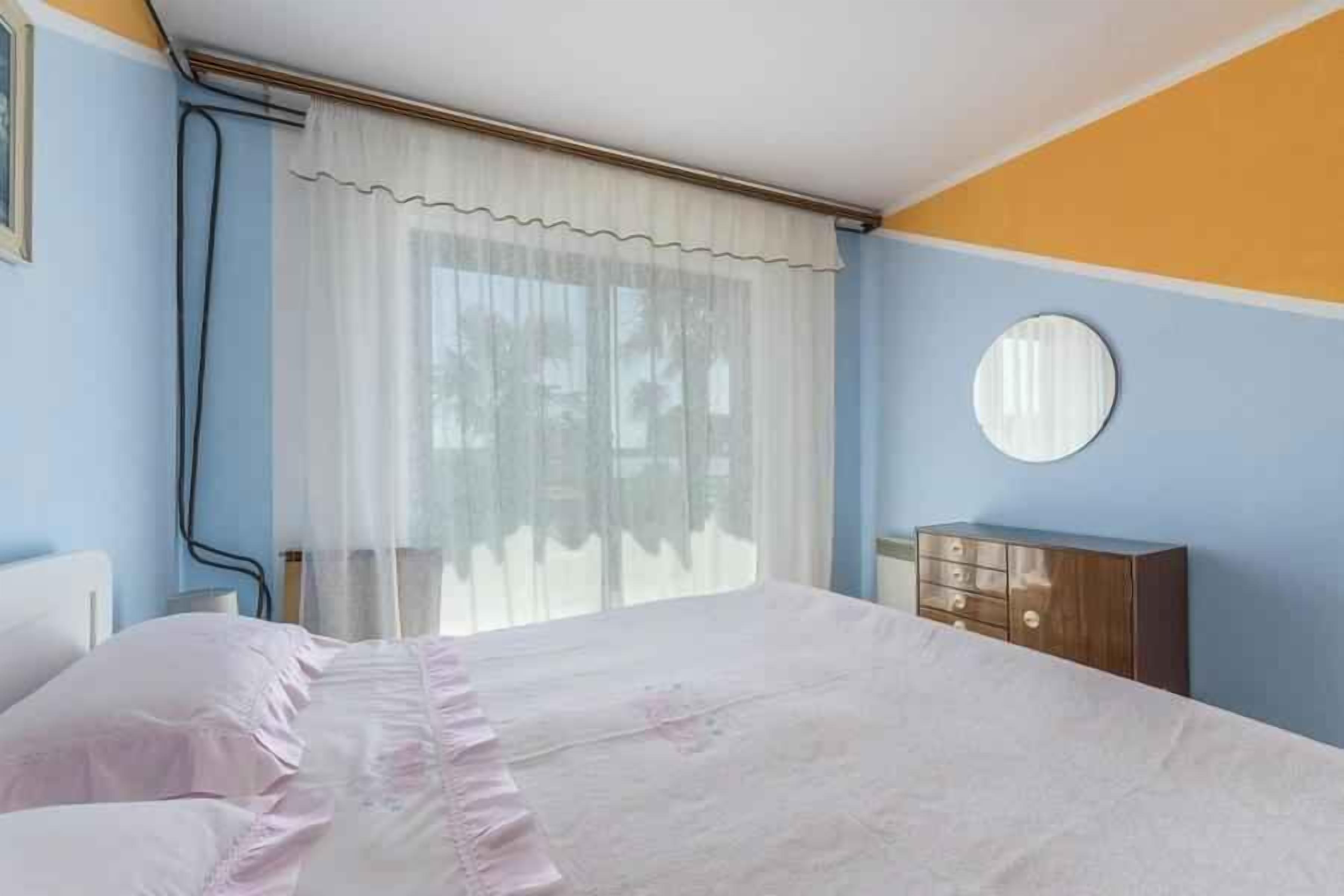 A bedroom with a bed, a dresser, a window with curtains, and a round mirror on the wall.