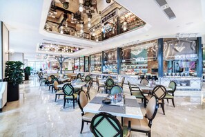 Een ruime, overdekte eethoek met meerdere tafels en stoelen, een hoog plafond met industriële verlichting en een uitzicht op het restaurantgedeelte door grote ramen.