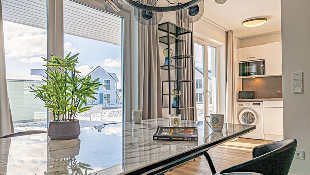 Ett modernt kök med tvättmaskin, en matplats med ett glasbord och utsikt över ett hus utanför.
