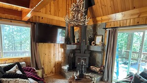 Ein gemütliches Blockhäuschen im Inneren mit Kamin aus Stein, Holzwänden, einem großen Fernseher und Blick ins Freie.