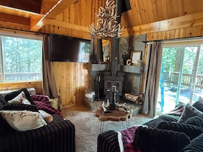 A cozy cabin interior with a stone fireplace, wooden walls, a large TV, and a view of the outdoors.