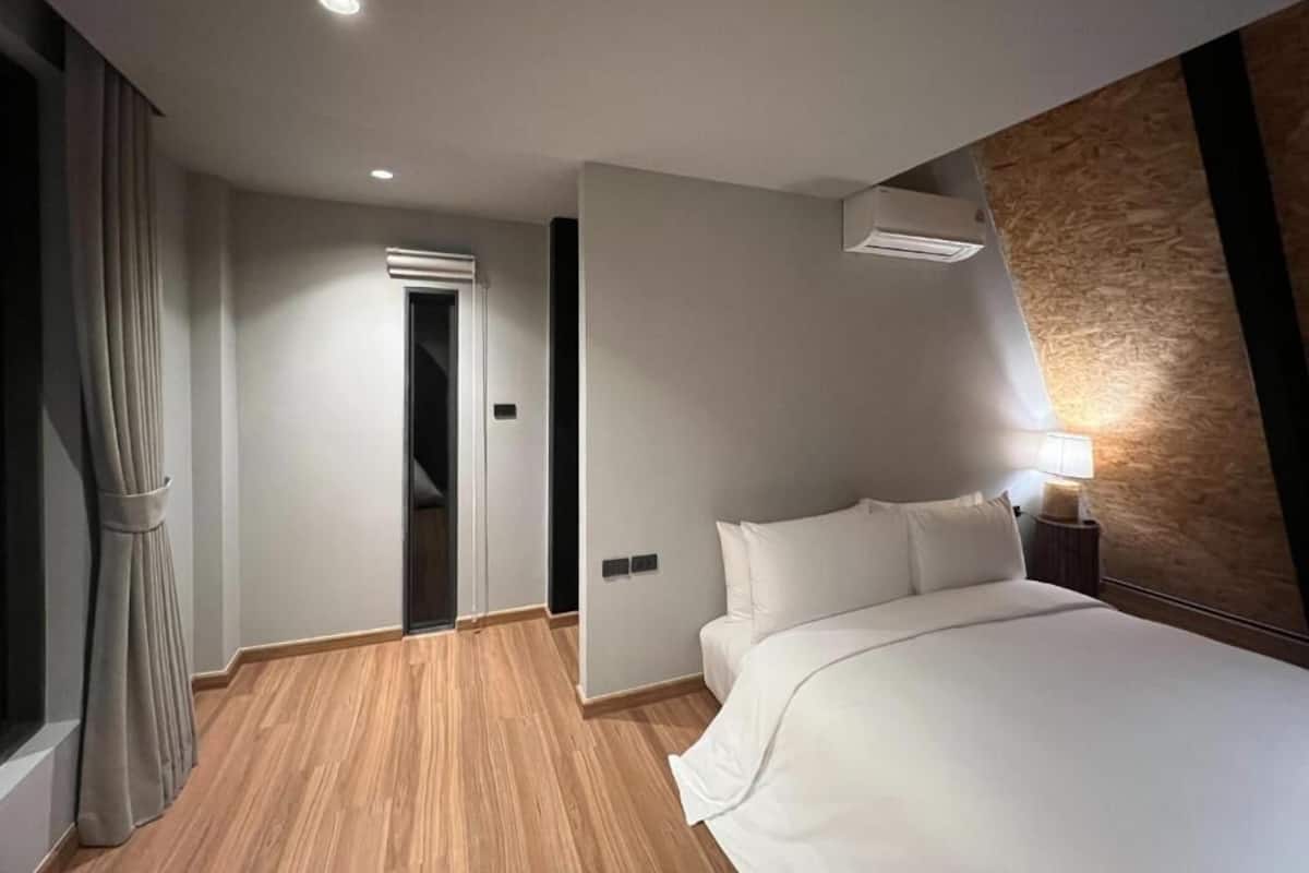A hotel room with a bed, a nightstand, a lamp, a window with curtains, an air conditioning unit, and a wooden floor.