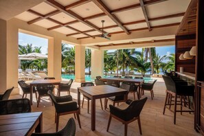 Une terrasse extérieure avec des tables et des chaises en bois, une piscine et des palmiers en arrière-plan.