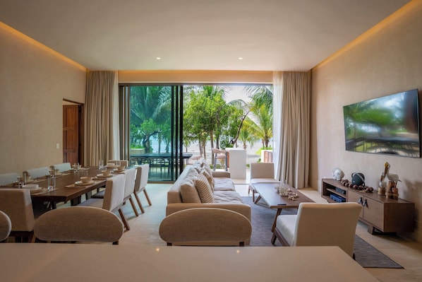 Une chambre d’hôtel moderne dotée d’un coin repas, d’un ensemble de canapés, d’une télévision et d’une grande fenêtre donnant sur un paysage verdoyant.