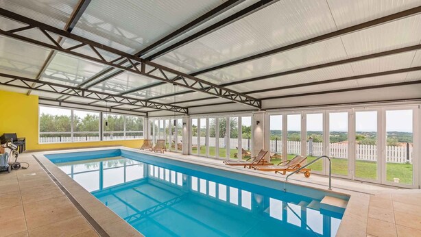 An indoor swimming pool with clear glass walls, a yellow wall on one side, and a view of a white picket fence and greenery outside. - Holiday apartment mit Klimaanlage und Pool (Umag)