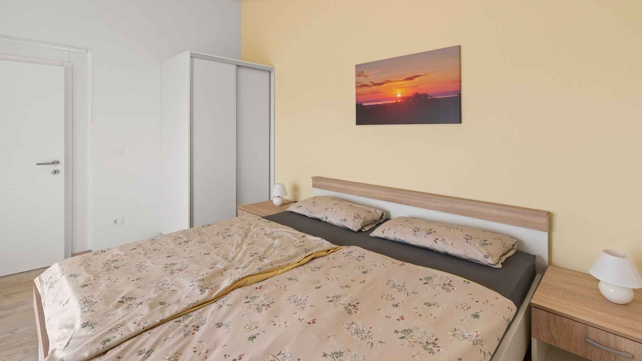 A bedroom with a bed, nightstands, a lamp, a wardrobe, and a sunset painting on the wall.