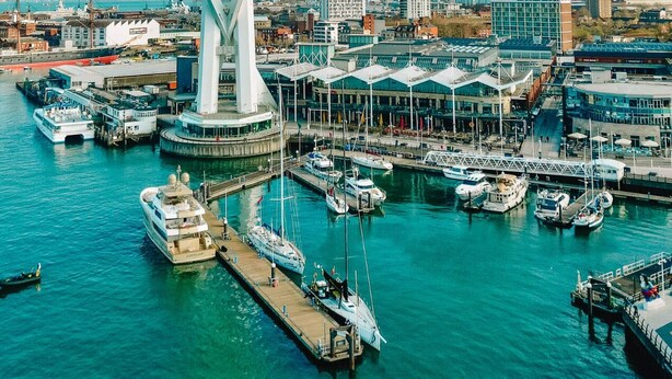 Una estructura arquitectónica moderna de diseño circular, rodeada por un puerto deportivo con barcos y un paisaje urbano al fondo.