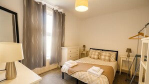Un dormitorio con cama, mesitas de noche, lámpara, radiador y ventana con cortinas.