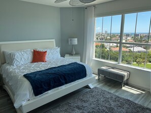 Ein Schlafzimmer mit Bett, Nachttisch, Lampe, einer Bank und einem großen Fenster mit Blick auf Gebäude und Bäume.