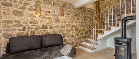 A stone-walled room with a sofa, a table with a book, a black wood-burning stove, and a staircase.