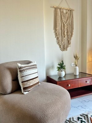 Ein Wohnzimmer mit einer Couch, einem Wandbehang aus Makramee, einem kleinen Tisch mit einer Vase und einer Pflanze.
