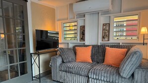 En stue med en grå polstret sofa, et sort fjernsyn på et stativ og et vindue med skodder.
