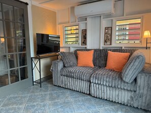 A living room with a grey upholstered sofa, a black TV on a stand, and a window with blinds. - NewReno, AC, 2 blocks to Beach, Dive, Eat, Drink (Frederiksted)