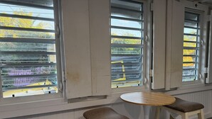 Et værelse med to barstole og et lille rundt bord, store vinduer med persienner, og udsigt til en gul varebil udenfor.