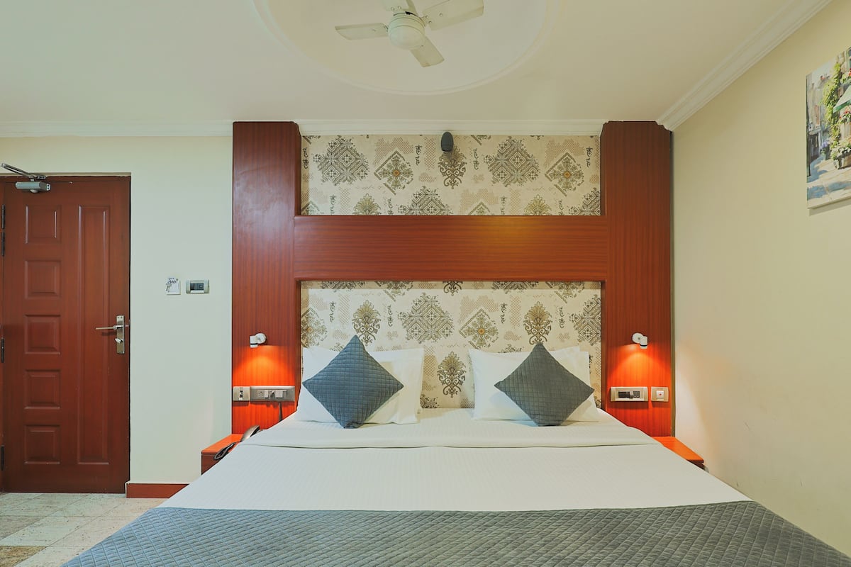 Habitación de hotel con cama, ventilador de techo, cabecero de madera, papel tapiz estampado e iluminación en la pared.