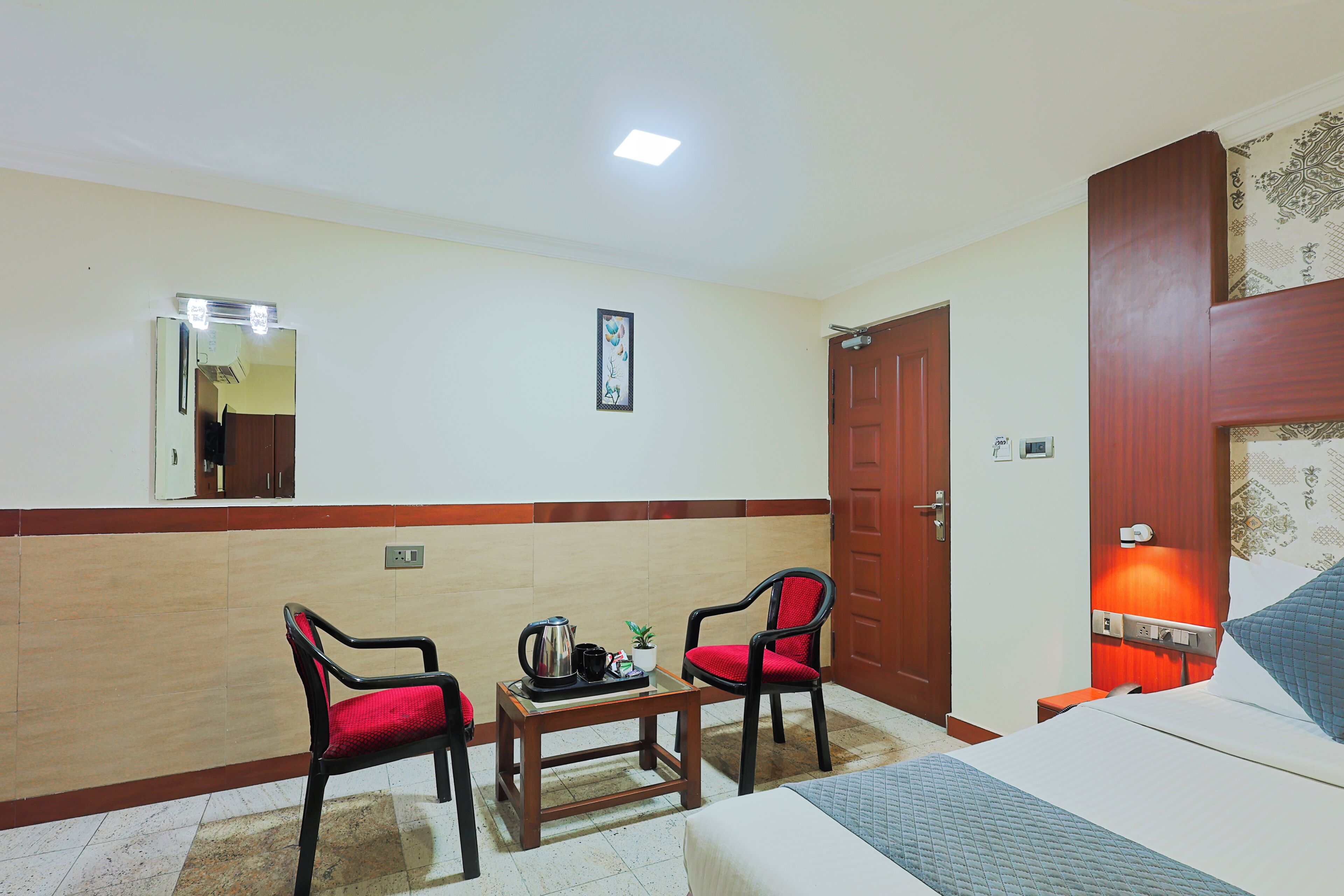 A hotel room with a bed, two chairs, a small table with a kettle and a plant, a mirror, a door, and a wall-mounted light.