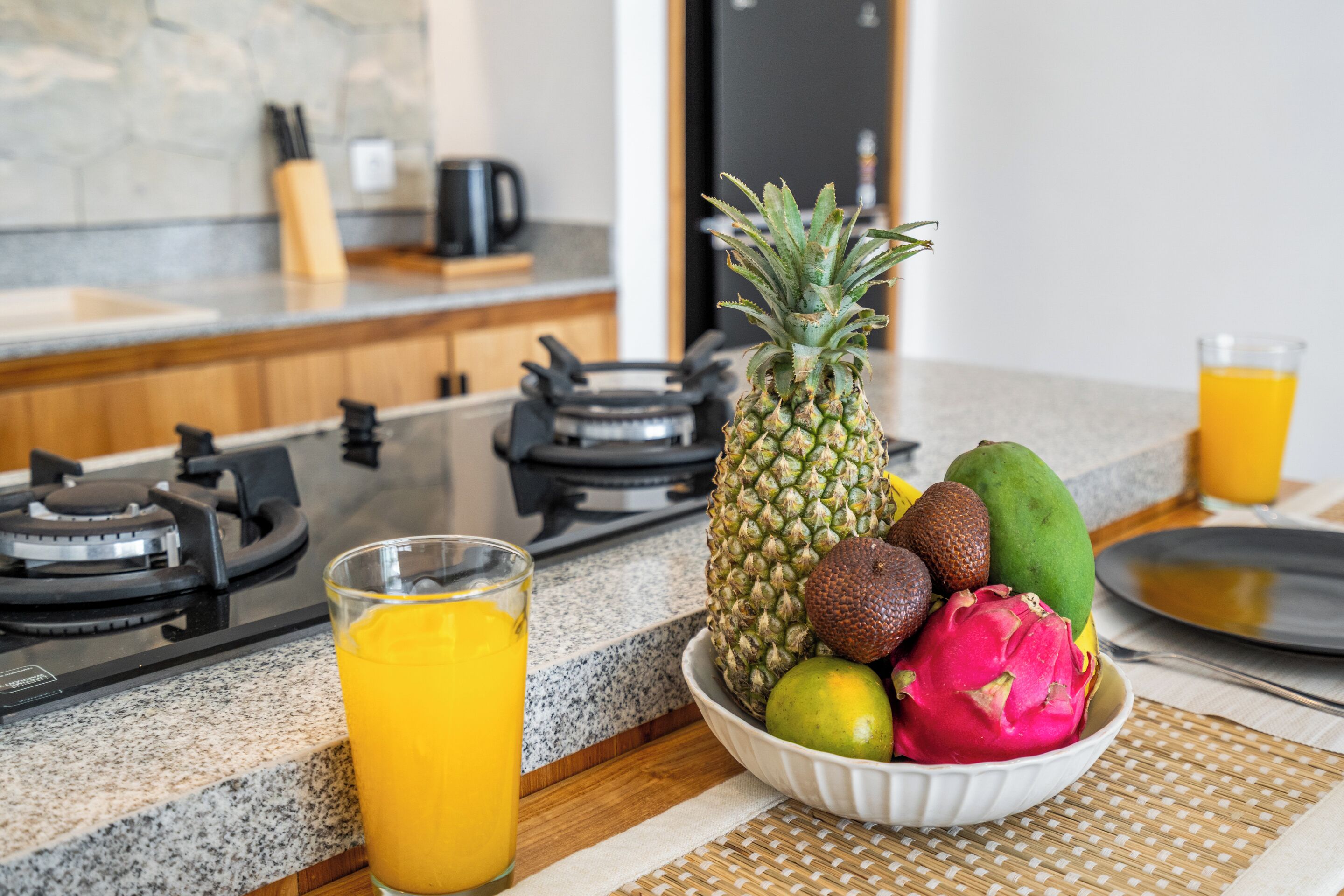 A kitchen counter with a fruit bowl containing pineapple, dragon fruit, and mango, a glass of orange juice, and a coffee maker in the background.