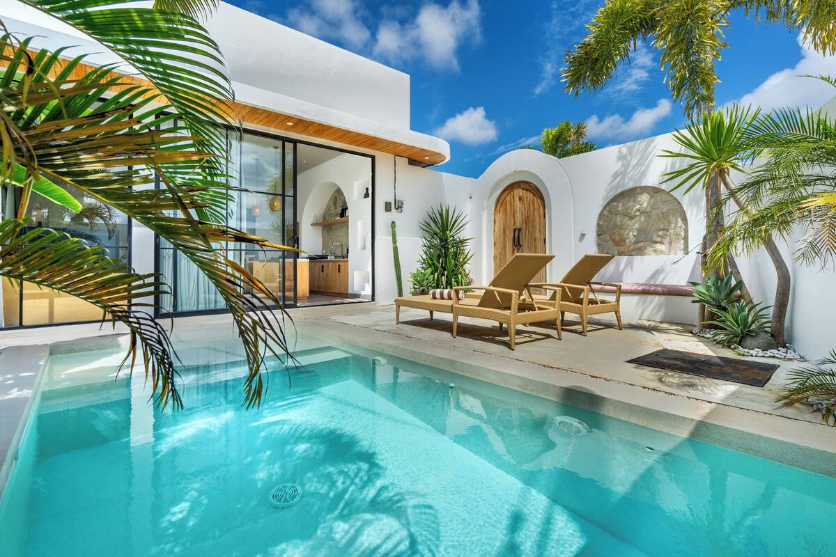 A modern pool area with a clear pool, wooden lounge chairs, and tropical plants.