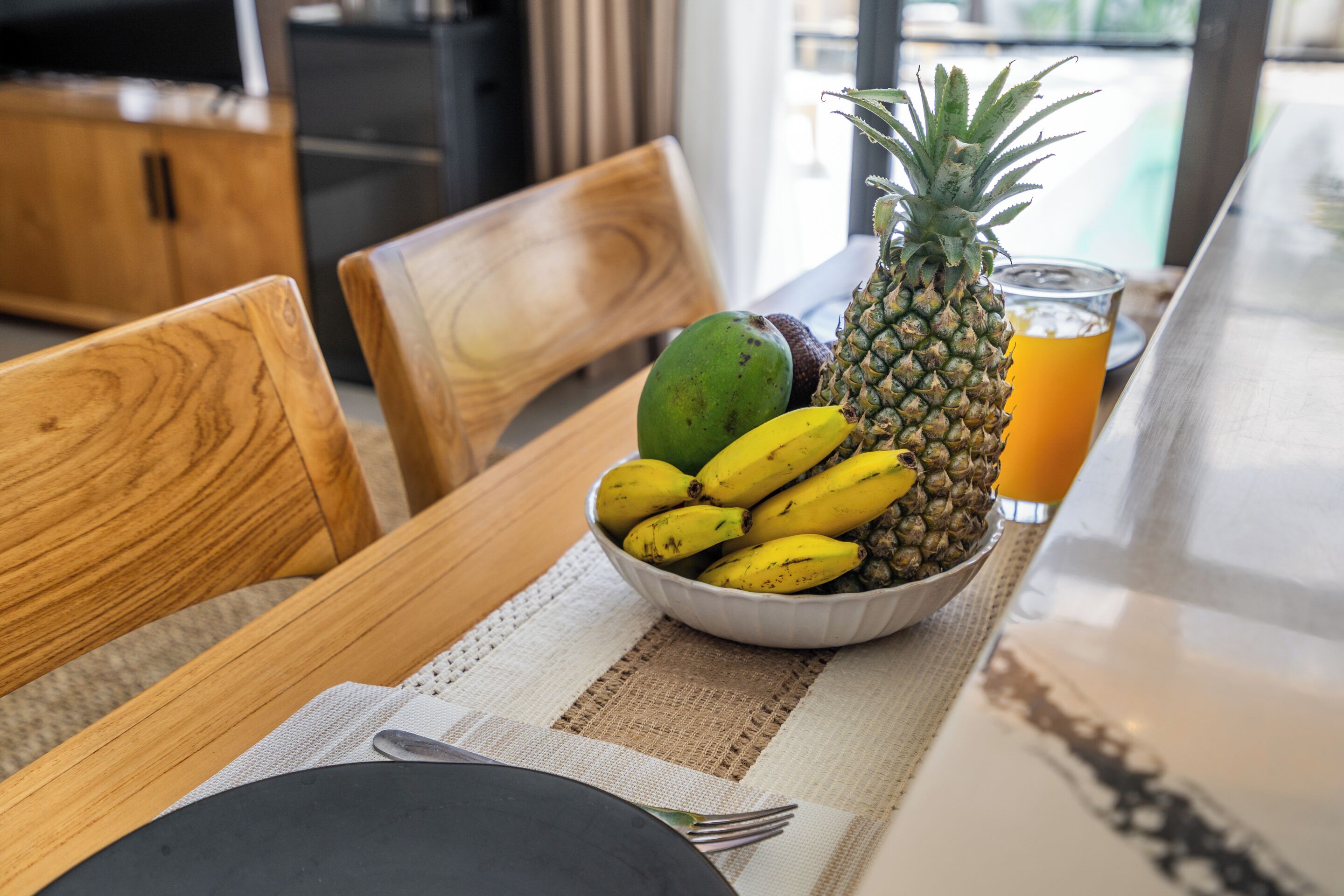 Une table à manger avec un saladier de fruits, un verre de jus d’orange et des chaises en bois.