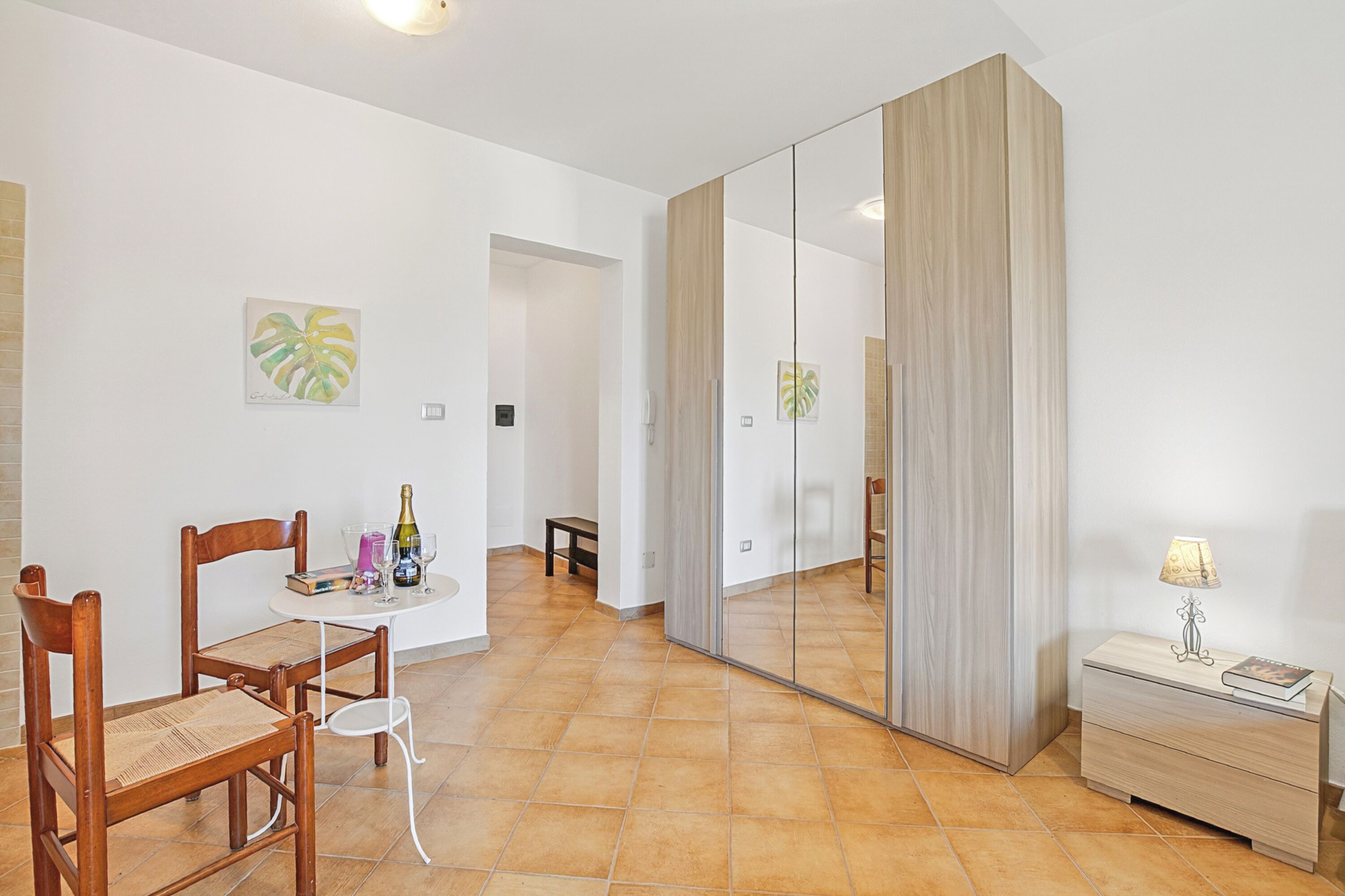 A room with a dining table, chairs, a mirror, a bedside table with a lamp, and artwork on the walls.