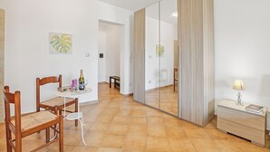 A room with a dining table, chairs, a mirror, a bedside table with a lamp, and artwork on the walls.