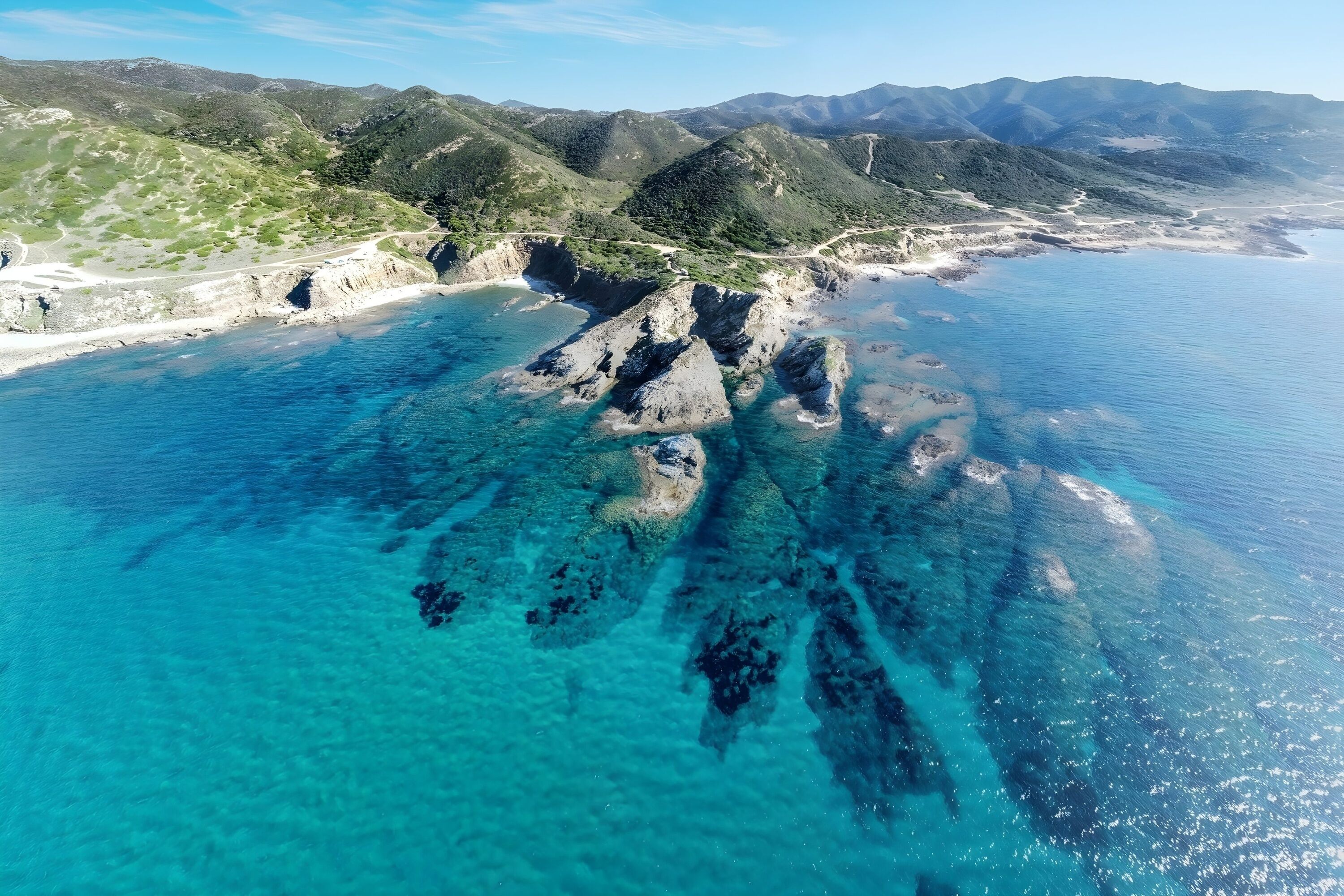 An aerial view of a coastal landscape with clear blue waters, rocky formations, and green hills.